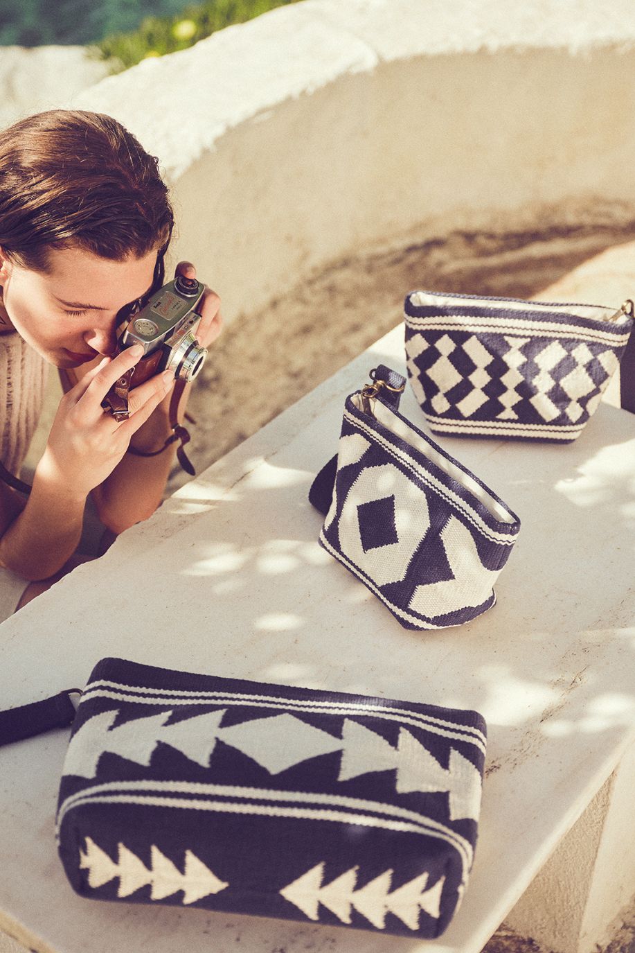 A woman leans over a table photographing a trio of woven Sea You Soon pouches, their geometric patterns arranged under dappled sunlight.