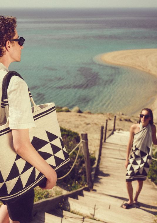 Man walking down a wooden path toward a calm beach with a geometric woven Sea You Soon tote bag, while a woman in a matching patterned pareo stands on the steps overlooking the sea.