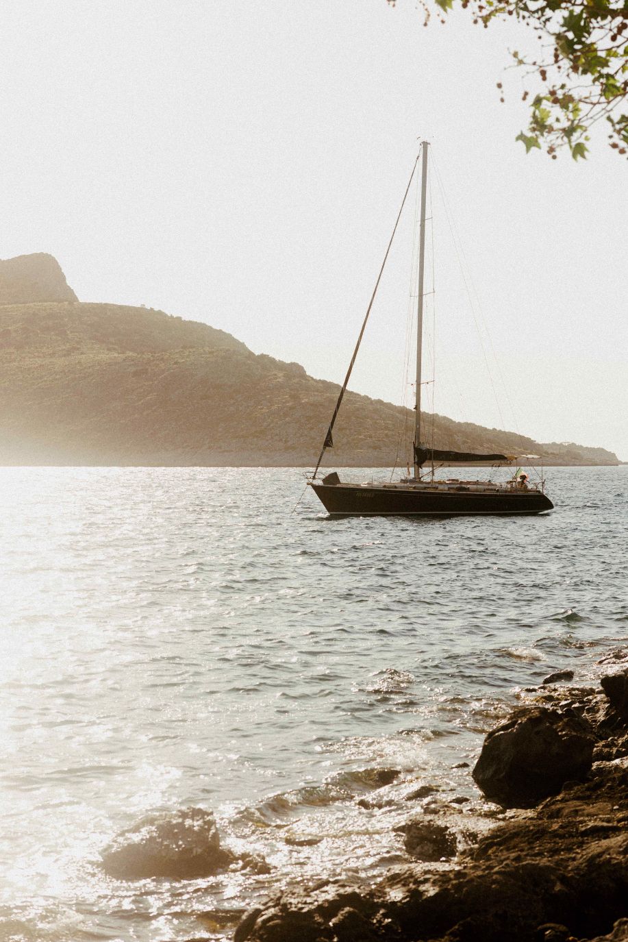 Sailboat anchored in a sunlit bay in Greece beside a rocky shore, with mountains in the background.