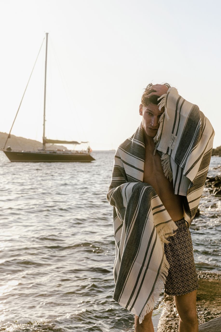 Man in patterned swim shorts wrapped in a striped beach towel beside the sea, with a sailboat behind him.