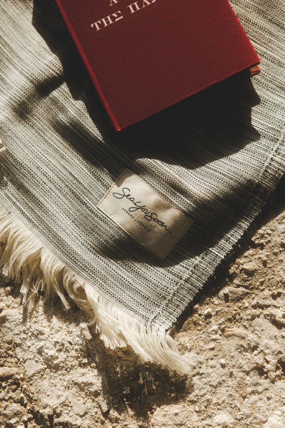 Close-up of a grey fringed lightweight beach towel with a Sea You Soon label and a red book resting on top.