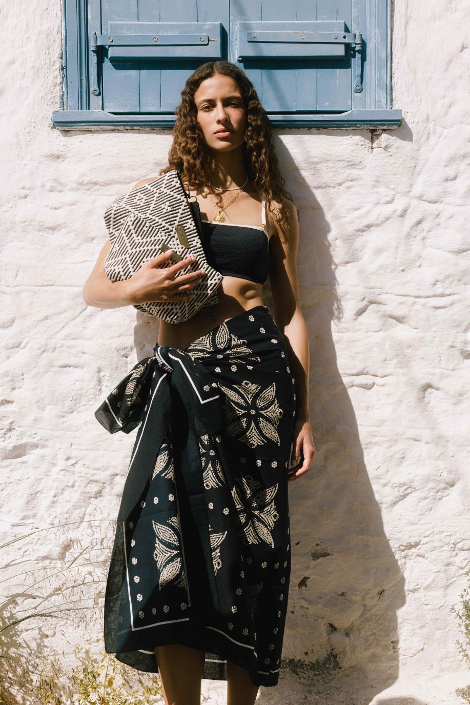 Woman wearing a black and cream pareo and holding a geometric clutch against a white wall beneath blue shutters.