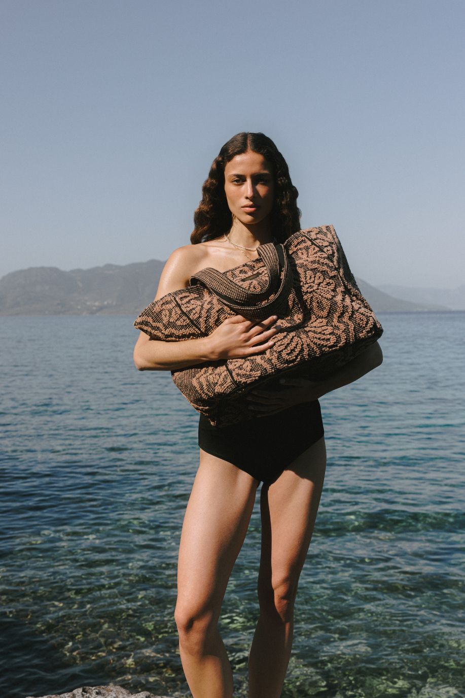 Woman in a black swimsuit holding a large brown and black patterned tote bag beside clear seawater.