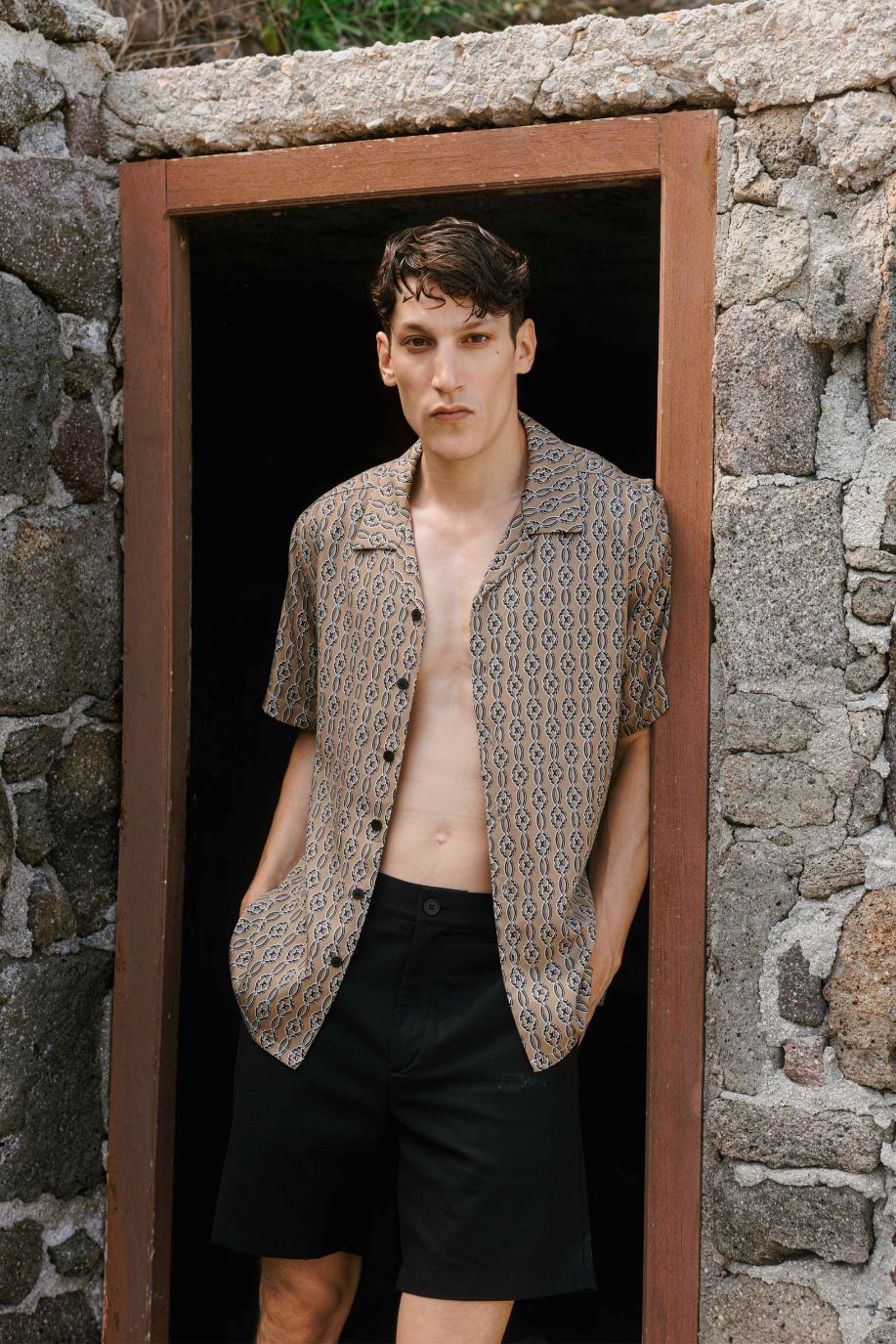 Man wearing an open patterned short-sleeved shirt and black shorts in a rustic stone doorway.